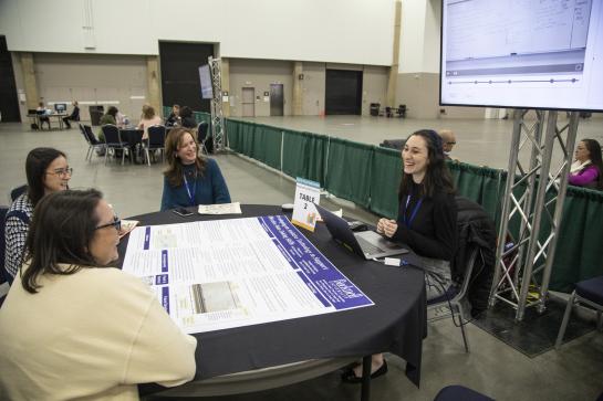 Four women sit talking at a round table. A large academic poster is on the table in front of them and a large TV behind them displays a presentation.